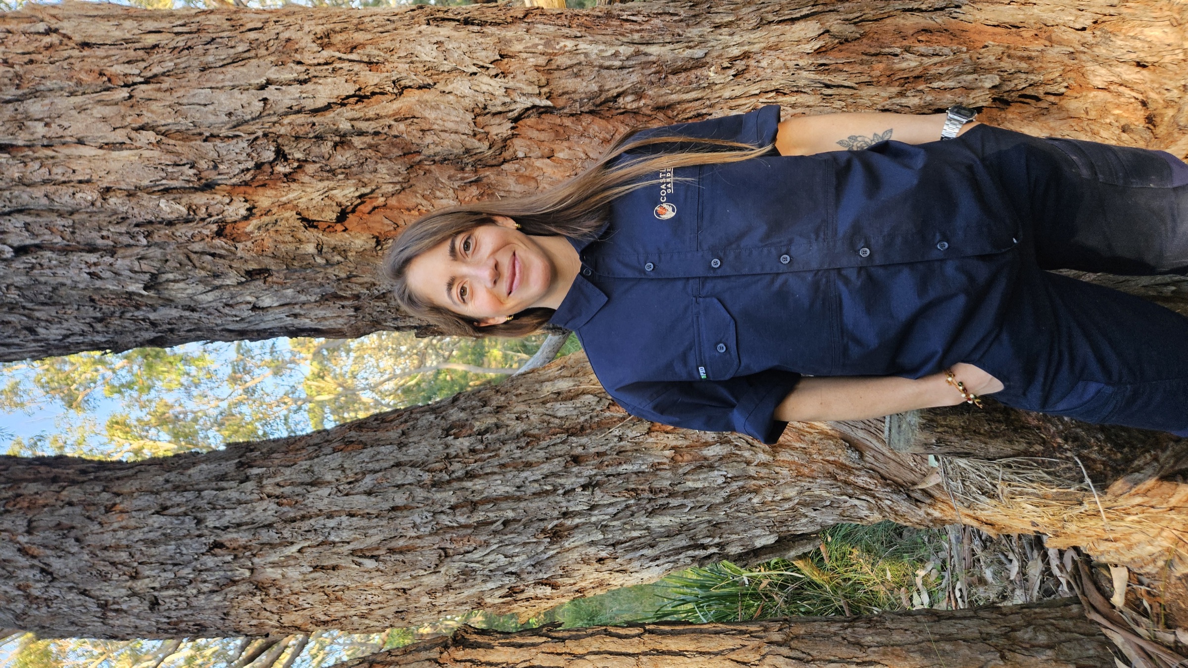 Mercedes wearing a Coastland Gardens shirt between two large gum trees